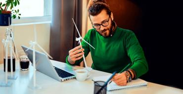 Hombre sentado frente al computador con maqueta de molino de viento tomando apuntes